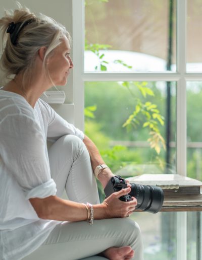 a woman looks out of a window in a sun filled room holding her camera during a personal branding photography shoot in surrey