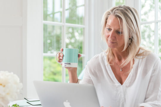 woman sits at her desk with a cup of coffee checking her computer