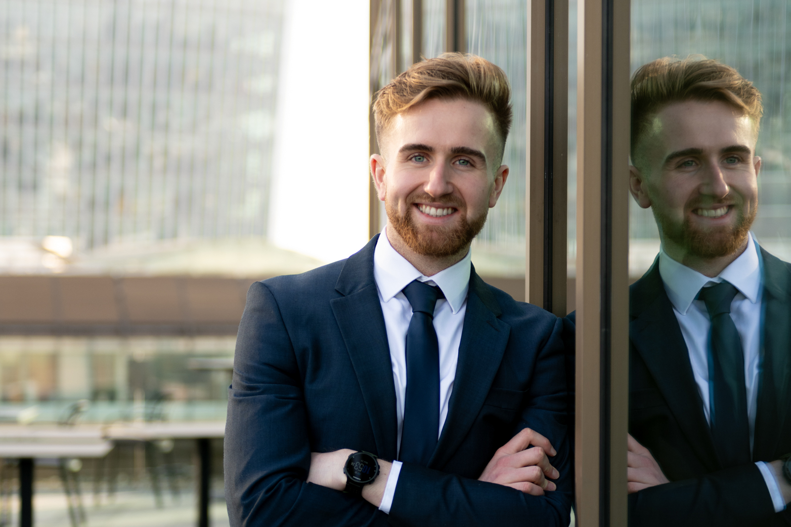 a young business man leans against a glass building in the city during a corporate headshot session