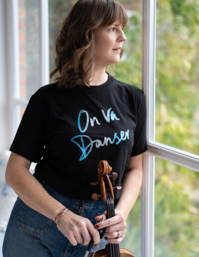 a woman poses at a large window with her violin