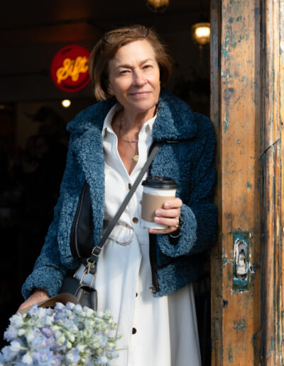 a woman holding a coffee cup and bunch of flowers leaning on a coffee shop doorway