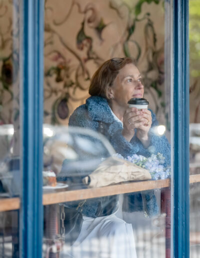 a woman holding a cup of coffee looks out of a coffee shop window during a personal brand photography shoot with surrey social stock photography