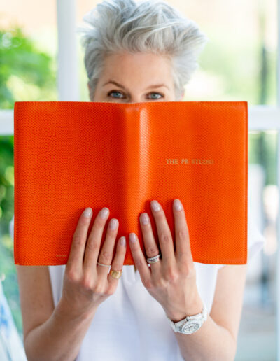 a silver haired woman peeks out from behind an orange book during a personal brand photo shoot in surrey